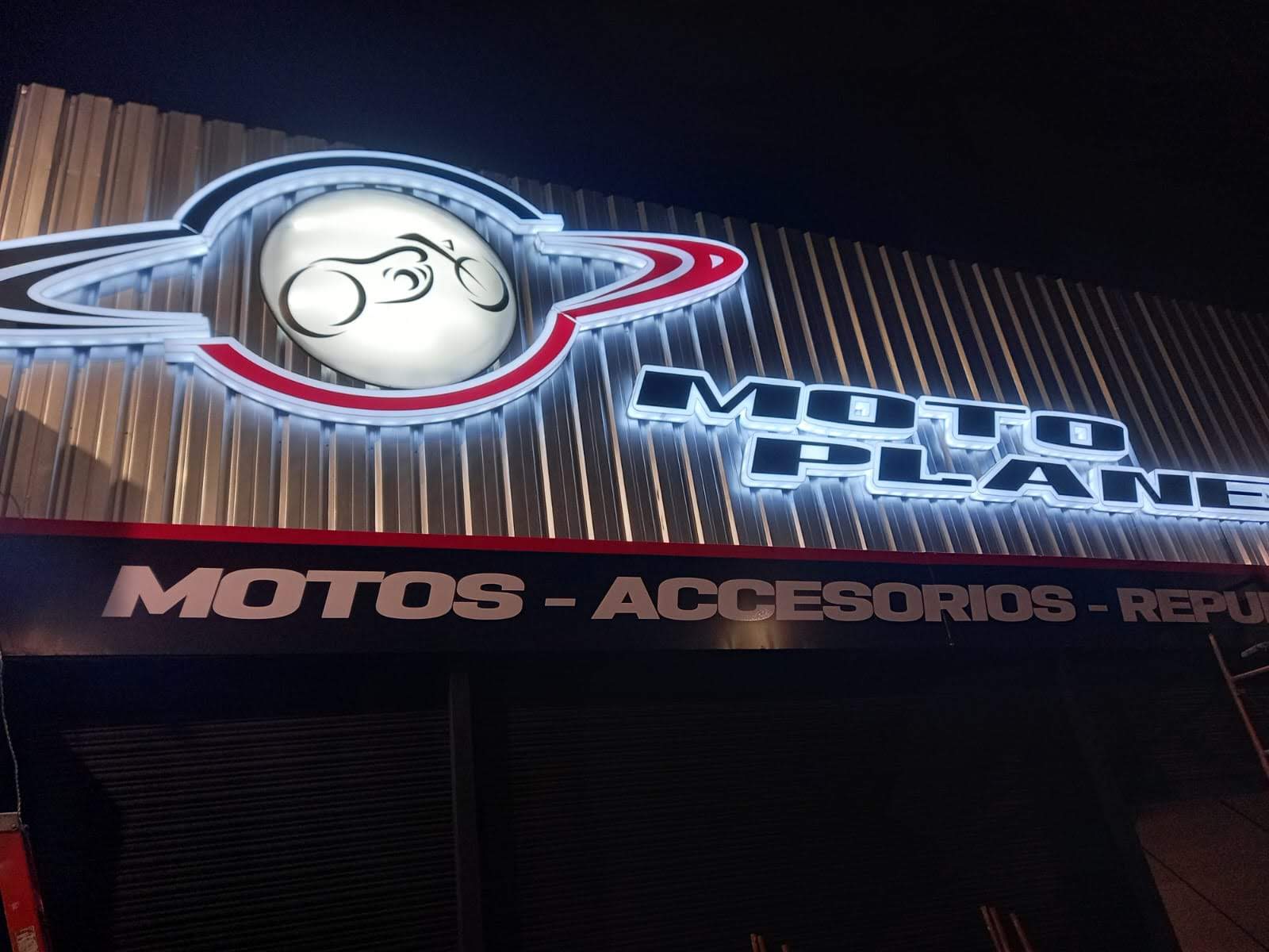 Fachada de Moto Planet de noche con logo y letras iluminadas sobre lámina metálica