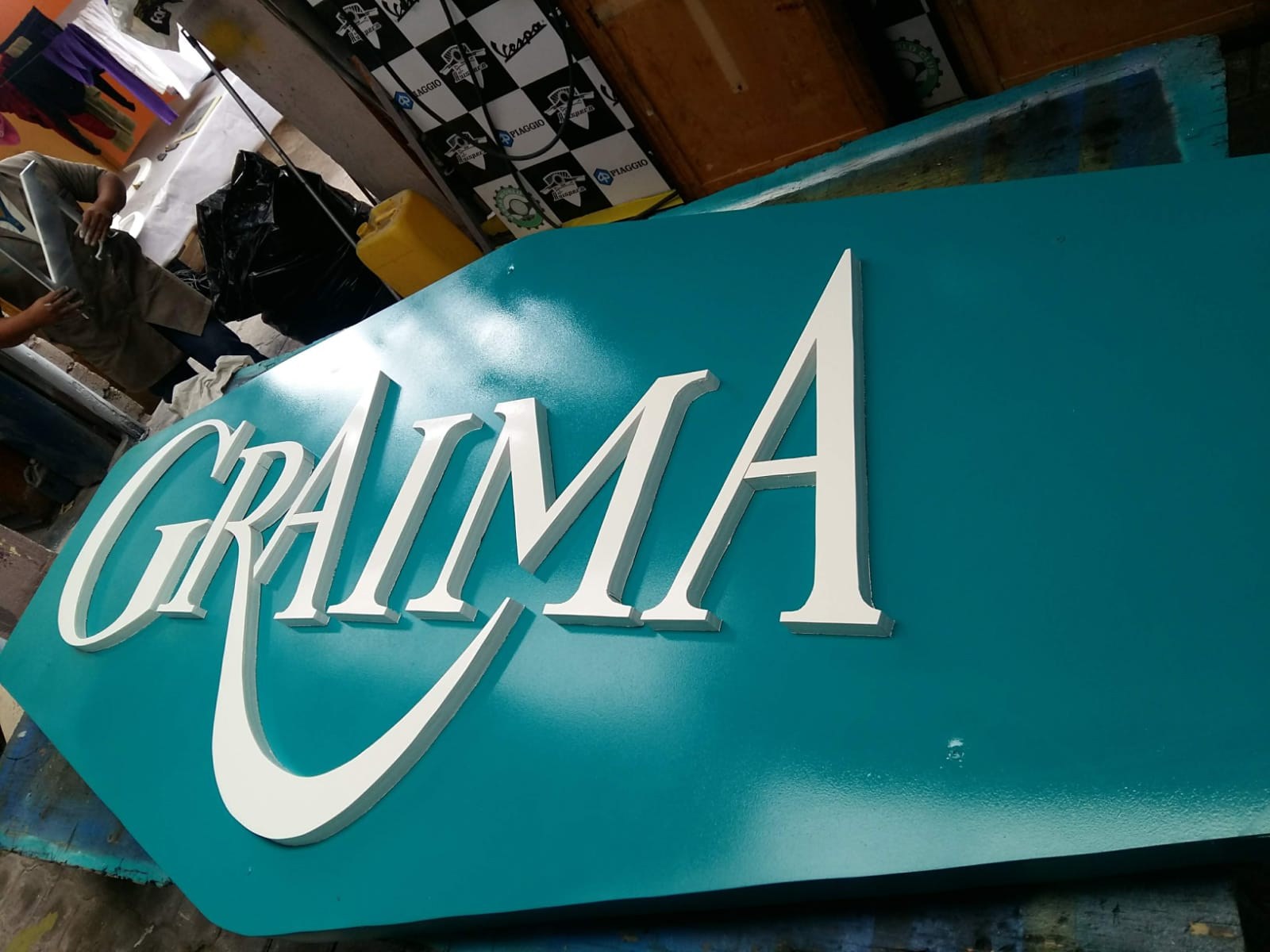 Letras corpóreas blancas de GRAIMA sobre un panel turquesa en el taller