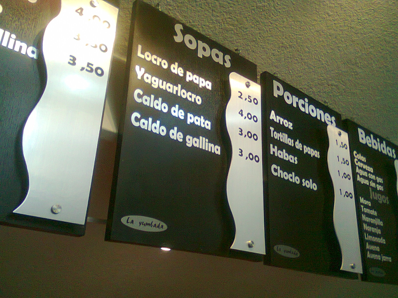 Menu boards negros de La Yumbada con sopas, porciones y bebidas, y precios sobre franjas de acero ondulado